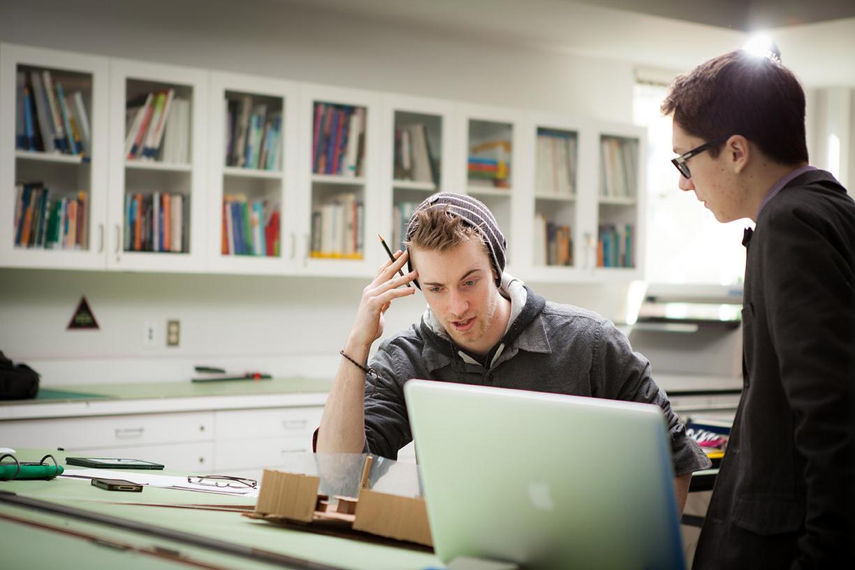 Student with headphones looking laptop screen with puzzled expression, another student looking at him