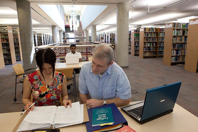A man and woman sit at a table in the library, looking at the woman's notebook.